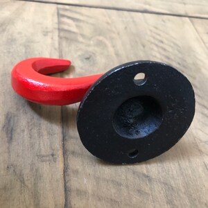 May include: A red metal hook with a black metal base. The hook is curved and has a smooth finish. The base has a round shape and a hole in the center for mounting.