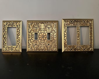May include: Three gold-colored decorative light switch covers with intricate floral designs. The covers are made of metal and have a polished finish.