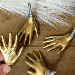 May include: Four gold-toned hand-shaped cabinet knobs with silver accents. The knobs are arranged in a circle on a wooden surface.