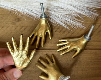 May include: Four gold-toned hand-shaped cabinet knobs with silver accents. The knobs are arranged in a circle on a wooden surface.