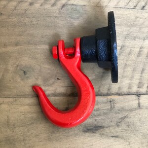 May include: A red metal hook with a black metal base. The hook is attached to the base with a screw. The hook is designed to be mounted on a wall.