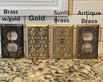 May include: Four decorative electrical outlet and switch covers in brass and gold tones. The covers are ornate with floral and scroll designs. The text above the covers reads "Brass w/gold", "Gold", "Sunlit Brass", and "Antique Brass".