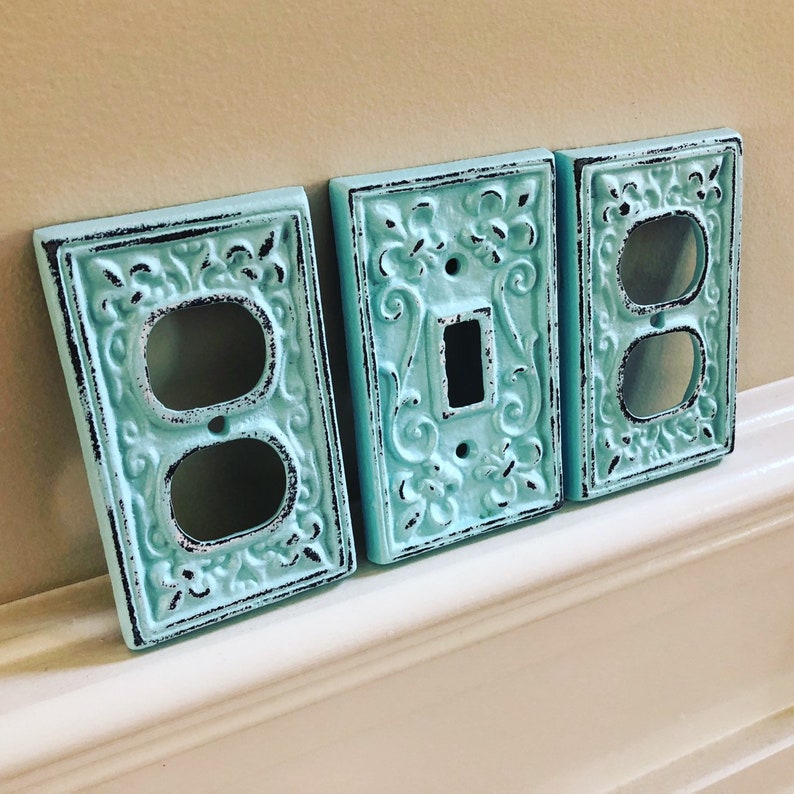 May include: Three decorative light switch covers in a light blue color with a distressed finish. The covers have an ornate design with a black border. The covers are for a single switch, a double switch, and a double outlet.