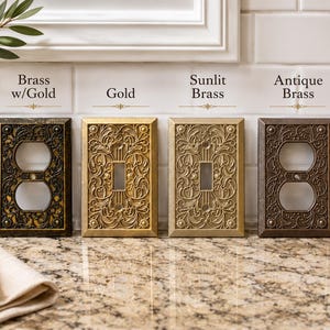 May include: Four rectangular wall plates with intricate floral scrollwork patterns. The finishes are labeled as Brass w/Gold, Gold, Sunlit Brass, and Antique Brass. Two plates feature a duplex outlet opening, and two feature a single toggle switch opening.