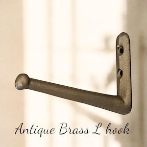 May include: An antique brass L-shaped hook with a rounded end and a mounting plate with two holes. The hook is a muted bronze color and appears to be made of metal. The text "Antique Brass L hook" is written in cursive below the hook.