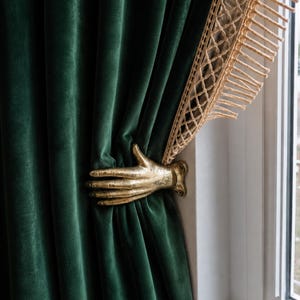 May include: A gold-colored metal hand-shaped curtain tieback holding a deep green velvet curtain in place. The curtain features a decorative beige macrame trim with fringe along the top edge, secured against a white window frame.