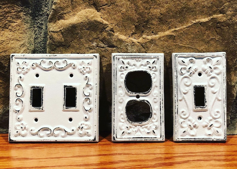 May include: Three white decorative light switch covers with ornate designs. The covers are made of cast iron and have a distressed finish.
