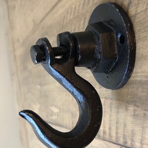 May include: Black metal hook with a round base and a screw. The hook is attached to a wooden surface.