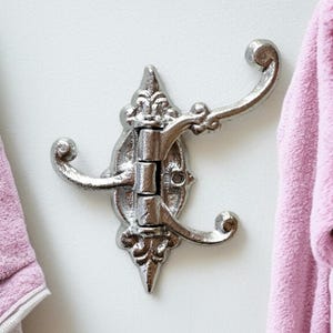 May include: Silver metal hook with a decorative design and three hooks. The hook is mounted on a white wall.