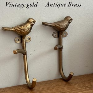 May include: Two decorative bird-shaped hooks in vintage gold and antique brass. The hooks are mounted on a wall and have a curved design. The text "Vintage gold" and "Antique Brass" is above the hooks.