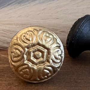 May include: Close-up of a gold-colored, round decorative knob with an embossed floral and geometric design. The knob is made of metal and has a slightly aged appearance. A black knob is partially visible in the background.
