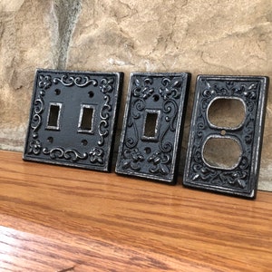 May include: Four decorative outlet covers in a black metal finish with an ornate design. The covers are for a double switch, a single switch, and a double outlet.