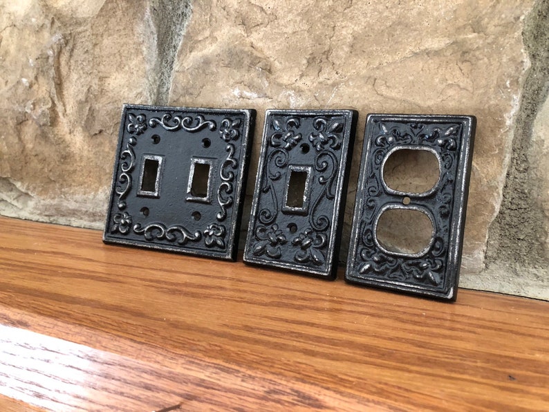 May include: Four decorative outlet covers in a black metal finish with an ornate design. The covers are for a double switch, a single switch, and a double outlet.