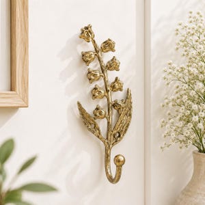 May include: A gold-toned metal wall hook shaped like a lily of the valley flower. The design features a central stem with multiple bell-shaped blossoms and two elongated leaves. The hook curves upward at the base and includes two visible screw holes.