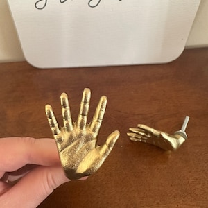 May include: Two gold-toned hand-shaped drawer pulls. One pull is a closed hand with five fingers, the other is an open hand with four fingers.
