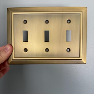 May include: A gold-toned, rectangular light switch plate with three toggle switches. The plate has a decorative border.