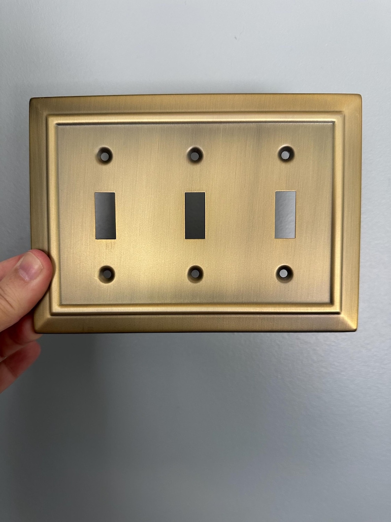 May include: A gold-toned, rectangular light switch plate with three toggle switches. The plate has a decorative border.