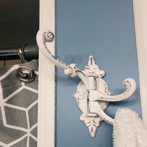 May include: White metal hook with a decorative design and three hooks. The hook is mounted on a blue wall.