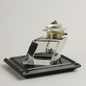 May include: A clear glass inkwell with a faceted lid and a black glass tray. The inkwell is shaped like a pyramid and has a small, clear glass bottle inside.