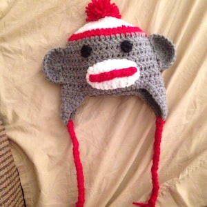 May include: A gray crocheted monkey hat with red and white stripes, a white pom-pom, and red yarn ties.