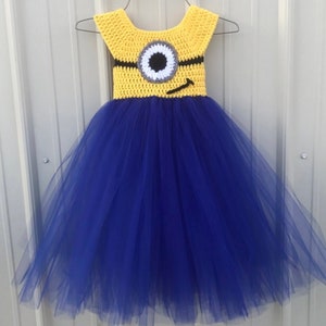 May include: A blue and yellow tutu dress with a crocheted Minion character on the bodice. The Minion has a single large eye, a black mouth, and a black strap across its head.