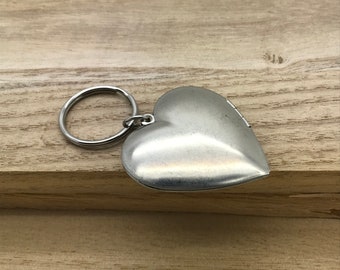 May include: A silver heart-shaped locket keychain with a ring attached. The locket is on a wooden surface.