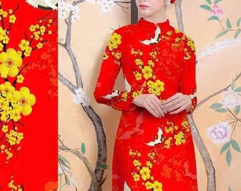 May include: A red dress with a floral pattern featuring yellow flowers and white birds. The dress has long sleeves and a high neckline. The dress is worn by a person with black hair and a headband. The background is a painted mural of a tree with white flowers.
