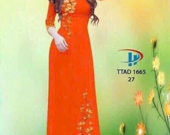 May include: A woman wearing a long, orange dress with floral embroidery. The dress has a fitted bodice and a flowing skirt. The woman is standing in front of a green background with flowers.