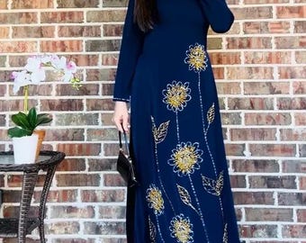 May include: A long, navy blue dress with gold floral embroidery. The dress features long sleeves and a beaded neckline. A small black purse is held in the hand. The background is a brick wall.