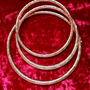 May include: Four gold-colored, intricately patterned necklaces arranged in a nested configuration on a red velvet surface. Each necklace features a detailed, repeating design along its circumference, creating a layered effect.