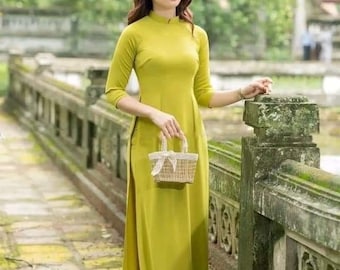 May include: A woman in a long, chartreuse dress with three-quarter sleeves and a mandarin collar. She holds a small woven basket with a white ribbon. The dress has a high slit. She wears a delicate headband and stands near a stone bridge.