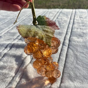 May include: A decorative cluster of orange-colored glass grapes with a green and red leaf. The grapes are faceted, and the stem is wrapped in green material. A gold string is attached for hanging. The grapes are held by a hand.