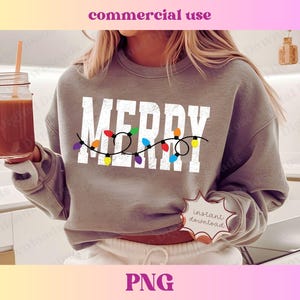 Puede incluir: Una sudadera gris con la palabra "Merry" en letras blancas con luces de cadena de colores alrededor. La sudadera la lleva puesta una persona que sostiene un vaso de líquido marrón.