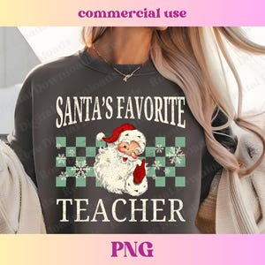 Puede incluir: Una sudadera gris con el texto "SANTA'S FAVORITE TEACHER" en blanco con un aspecto vintage desgastado. Una imagen de dibujos animados de Pap&aacute; Noel con un gorro rojo y barba blanca est&aacute; en el centro del texto.