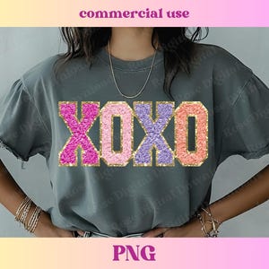 Puede incluir: Una camiseta gris con un gráfico brillante de "XOXO" en rosa, morado y dorado.
