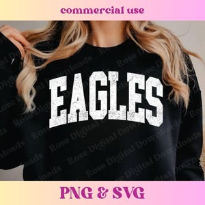 以下が含まれることがあります： 白地に「EAGLES」とヴィンテージ風のダメージ加工が施された黒いスウェットシャツ。長いブロンドの髪をした人が着用しています。