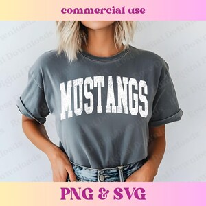 Pu&ograve; includere: Una maglietta grigia con la parola "MUSTANGS" stampata in bianco sul davanti. La maglietta &egrave; indossata da una persona con i capelli lunghi e biondi.