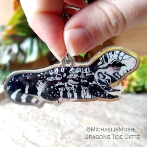 May include: A black and white lizard keychain with a clear acrylic background. The lizard is in a side profile with its mouth open. The keychain is attached to a silver metal ring. The text "@MICHAELISMOSHU DRAGONS TIDE GIFTS" is visible in the bottom right corner of the image.