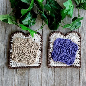 May include: Two handmade crochet coasters shaped like slices of toast, with a brown crust and a beige center. One coaster has a tan crochet design, the other a purple design. Green leaves are in the background.