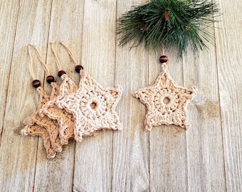 Puede incluir: Cinco estrellas de crochet beige con cuentas de madera colgando de un cordel. Los adornos están dispuestos sobre una superficie de madera marrón claro.