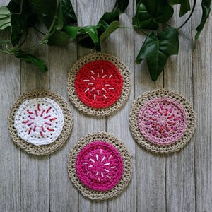 May include: Four handmade crochet coasters in shades of red, pink, and white, with a beige border. The coasters feature a circular design with radiating stitches, set against a wooden background with green foliage.