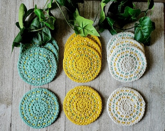 May include: Six crocheted coasters in three colors: teal, yellow, and cream. Each coaster has a white crocheted border and a center circle in a contrasting color.