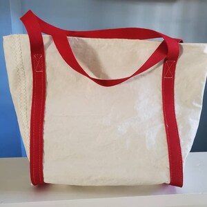 May include: A white tote bag with red handles and accents. The bag has a rectangular shape and is made of a textured material. The red handles are attached to the bag with red stitching.