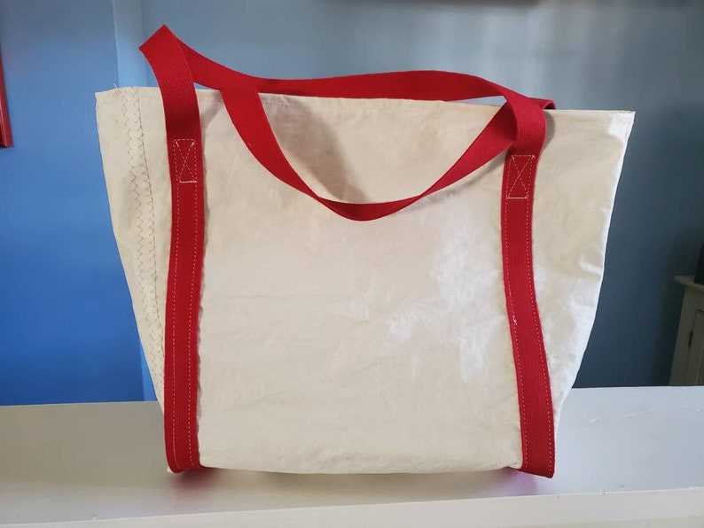 May include: A white tote bag with red handles and accents. The bag has a rectangular shape and is made of a textured material. The red handles are attached to the bag with red stitching.