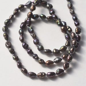 May include: A strand of dark, iridescent baroque pearls featuring irregular, organic shapes. The pearls exhibit deep purple, bronze, and charcoal tones with a high-luster finish, strung together on a thin, light-colored cord in a continuous loop.