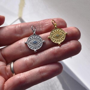 May include: Two silver and gold eye charms with a detailed design. The charms are round and have a raised eye design in the center. The eye is surrounded by a circle of small spikes. The charms are hanging from a chain.