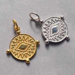 May include: Two silver and gold pendants with a stylized eye design. The pendants are round and have a textured surface. The eye design is in the center of each pendant and is surrounded by a circle of spikes.