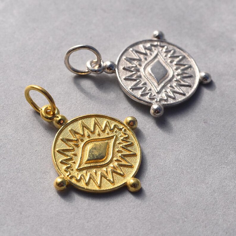 May include: Two gold and silver pendants with a stylized eye design. The pendants are round and have a textured surface. The eye design is in the center of the pendants and is surrounded by a circle of spikes.