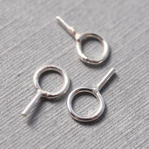 May include: Three small, silver-colored eye pins with a loop and a straight post. The eye pins are arranged on a gray surface. These findings are used in jewelry making and other crafts.