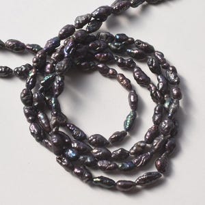 May include: A long strand of dark purple and black baroque pearls with an irregular, textured surface. The pearls exhibit a metallic luster with subtle iridescent highlights of blue and green, arranged in a continuous, coiled, multi-layered loop.